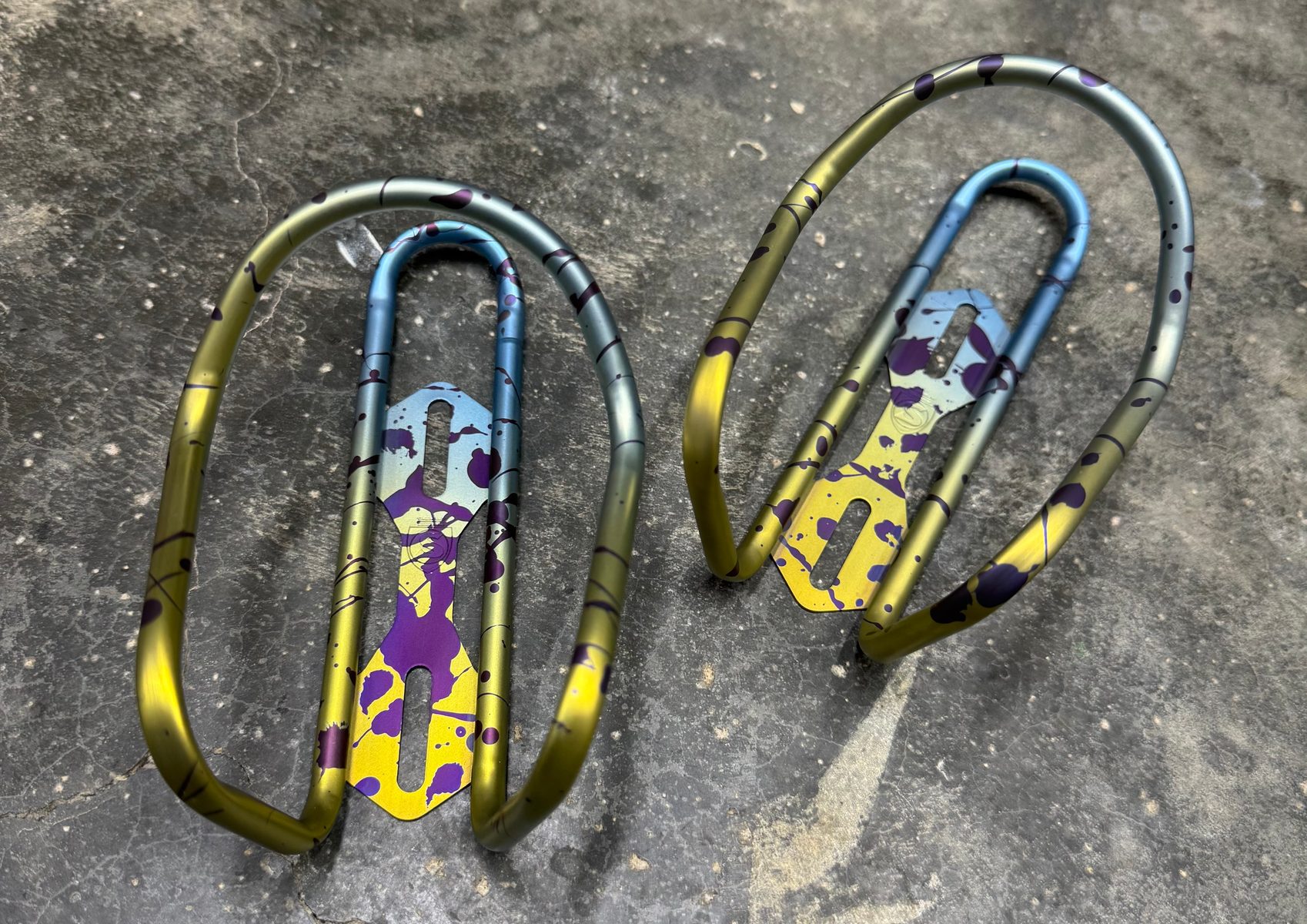 Pair of titanium bottle cages with a custom two-color gold-to-violet fade, shown with a matching stem cap and bolts