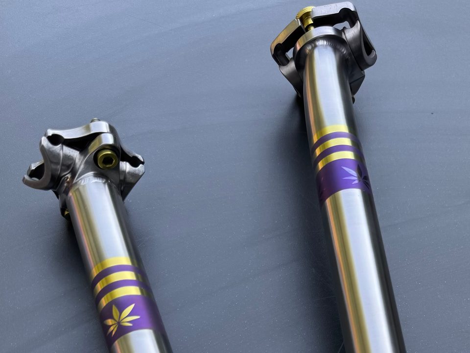 Two titanium seatposts with anodized gold bands and a gold leaf emblem on a violet collar