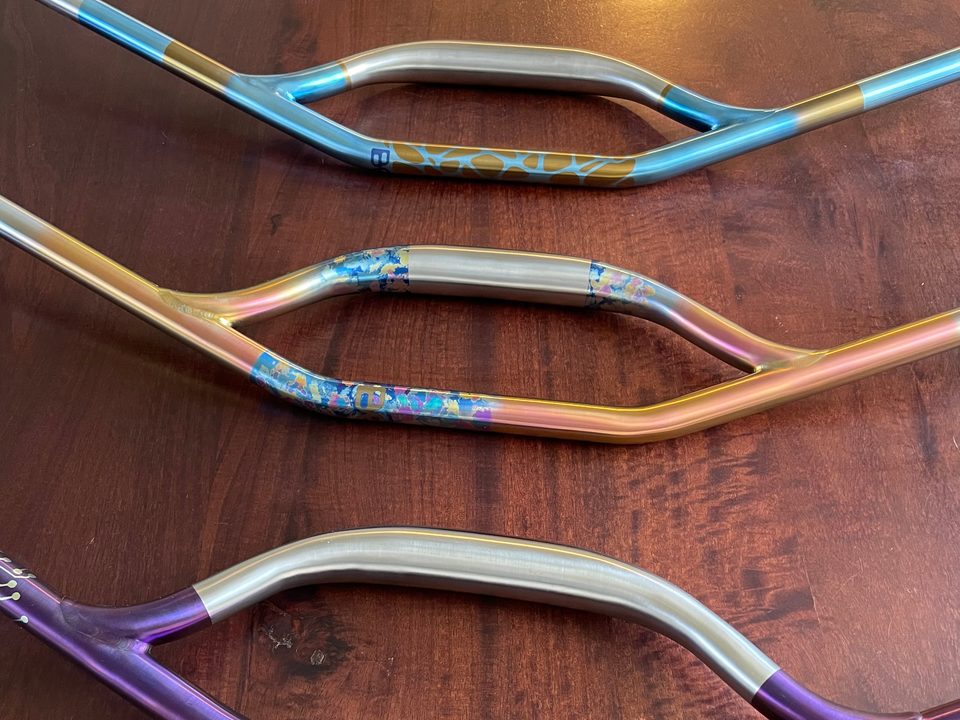 Three anodized titanium riser handlebars — blue and gold, pink and gold with a splatter section, and purple with a silver crown