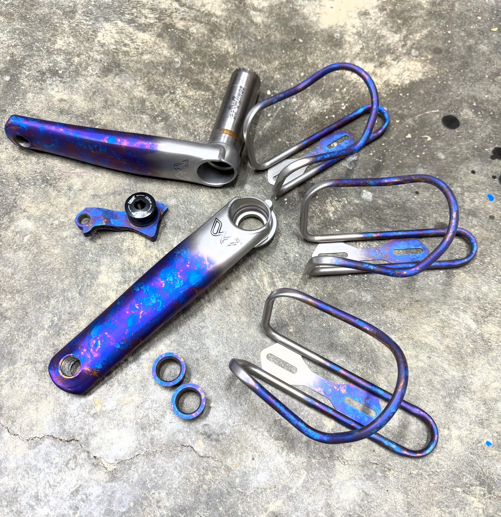 Anodized titanium crank arms, bottle cages and hardware laid out on concrete